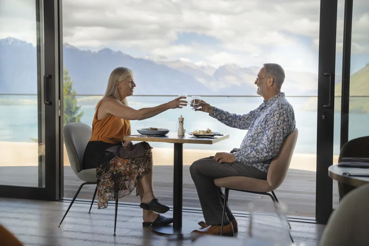 An elderly couple clinking their wine glasses together in celebration at a beautiful restaurant overlooking the beautiful waters of New Zealand.