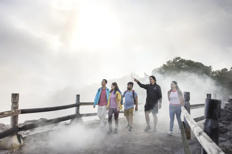 Group of people walking along a scenic path with private geothermal access in Rotorua Bay, New Zealand, surrounded by steam vents, natural hot pools, and lush greenery.
