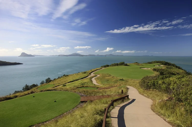 Scenic view of a golf course on a hillside overlooking a body of water with distant islands and a partly cloudy sky.