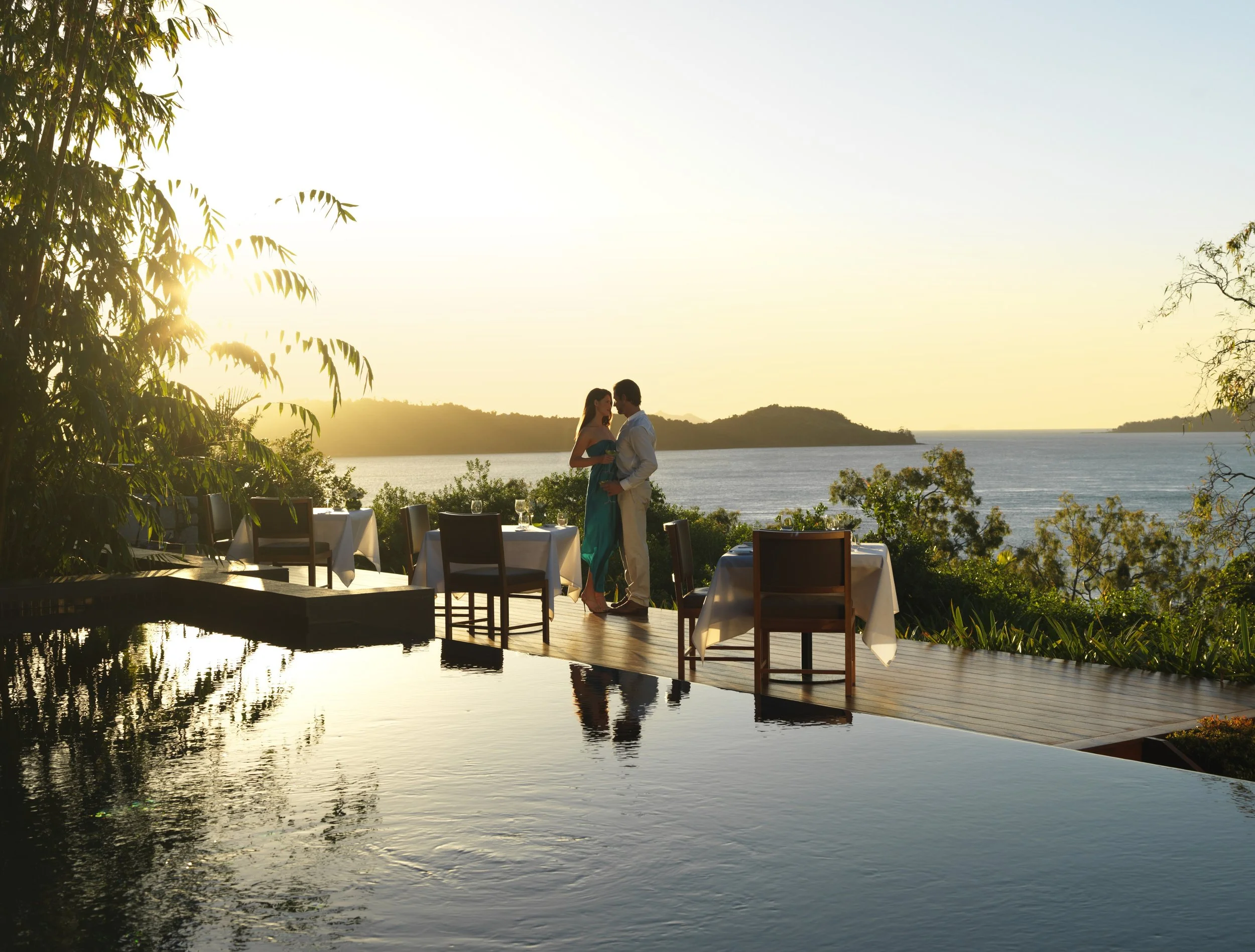 A couple embraces at sunset on a deck overlooking the ocean at qualia in the Great Barrier Reef, Australia, with dining tables, lush greenery, and their reflection in a calm pool creating a romantic, tranquil scene.
