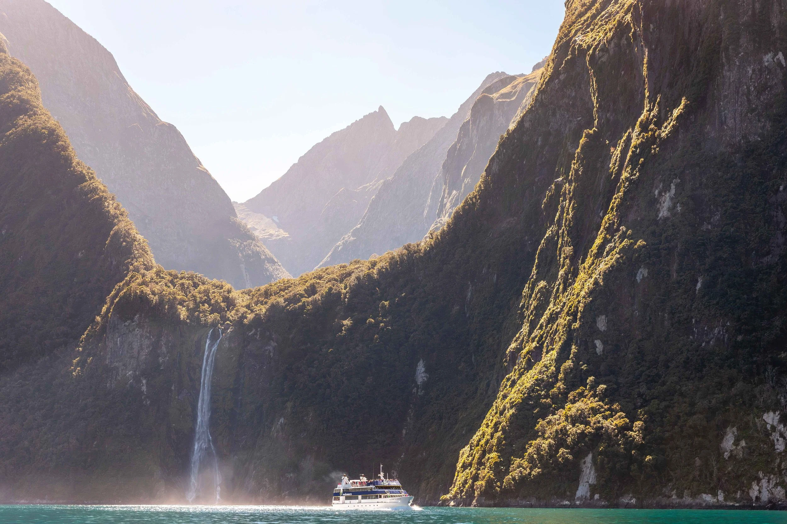 A boat cruises through Milford Sound in Fiordland, New Zealand, passing towering cliffs and cascading waterfalls in a dramatic fjord landscape