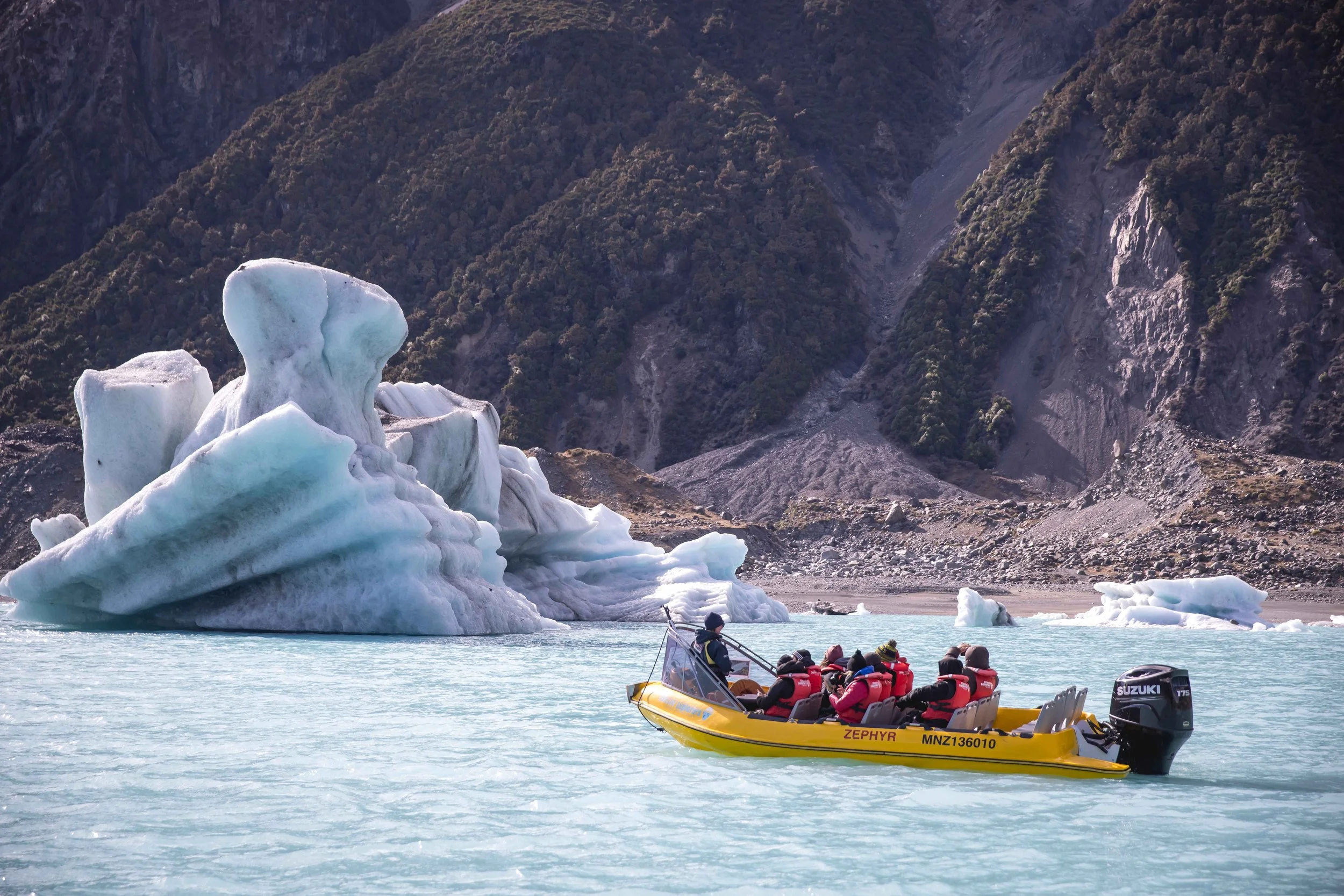 A boat navigates Tasman Lake among floating icebergs, with the snow-capped peaks of Aoraki/Mount Cook towering in the background, New Zealand