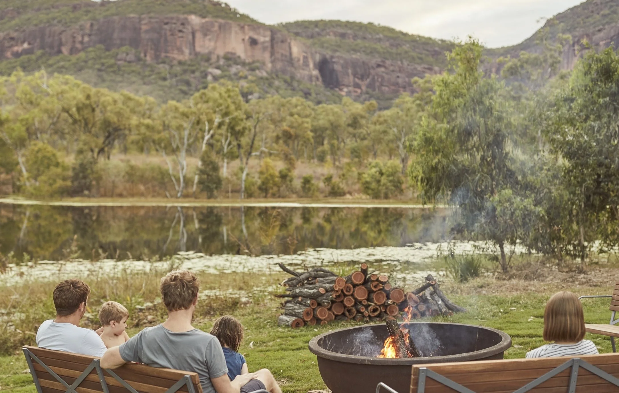 A family sits around a burning fire pit near a lake with a mountain in the background at Mt Mulligan Lodge in Northern Outback Queensland, Australia, enjoying a peaceful outdoor setting.