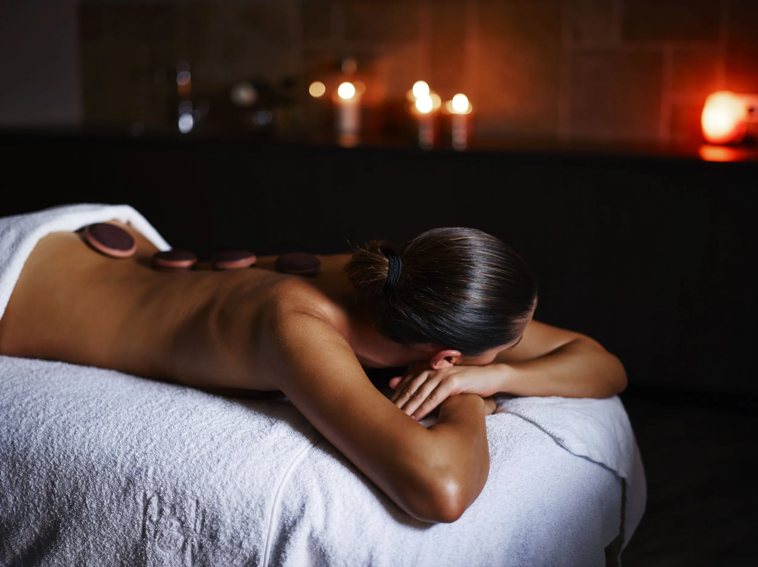A woman receives a hot stone massage at Pretty Beach House in New South Wales, Australia, lying face down on a white towel-covered table in a dim, candlelit spa setting.