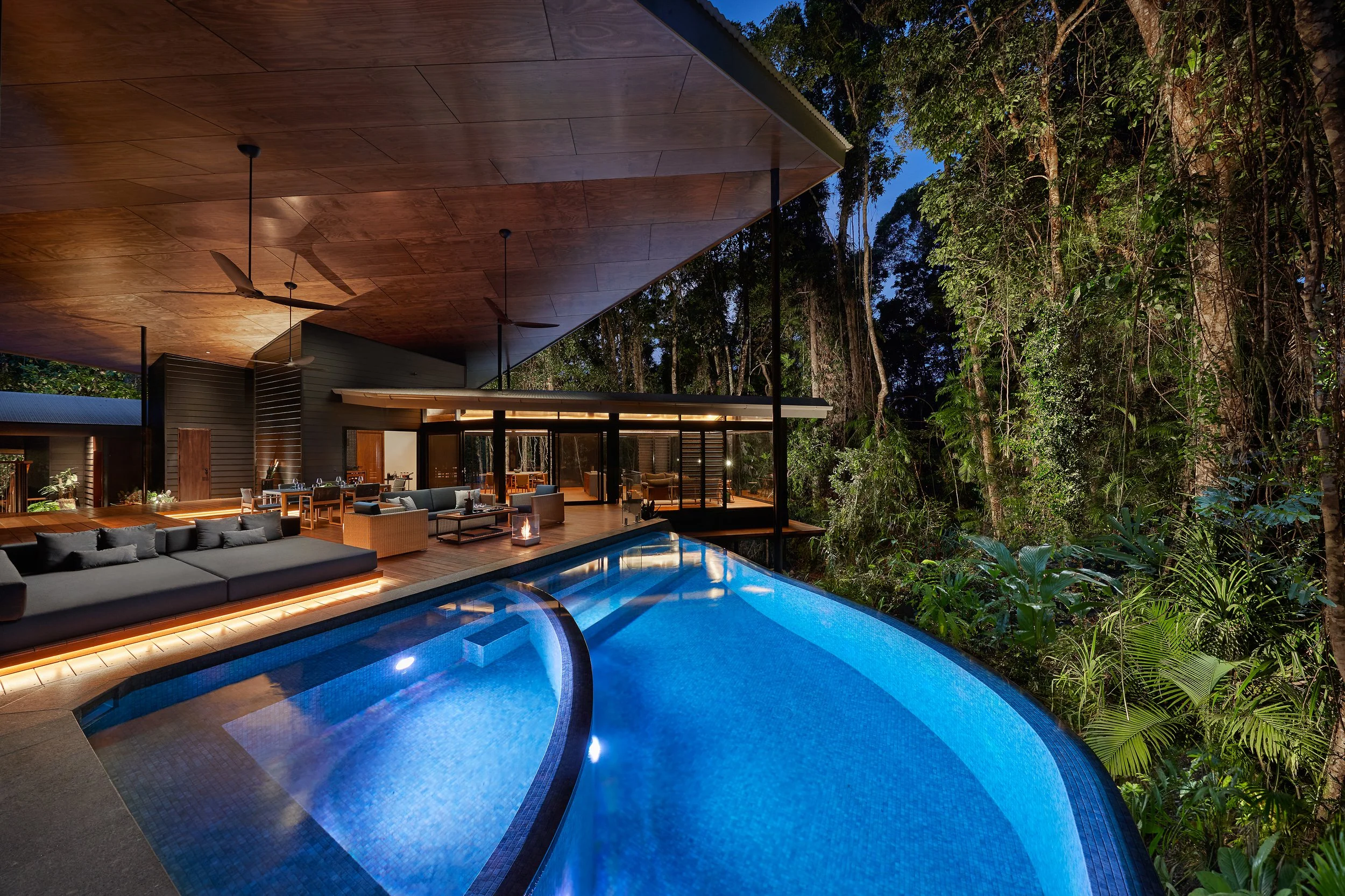 Outdoor pool area at Silky Oaks Lodge in Queensland, Australia, illuminated with blue lights, surrounded by lush greenery and modern treehouse-style architecture.