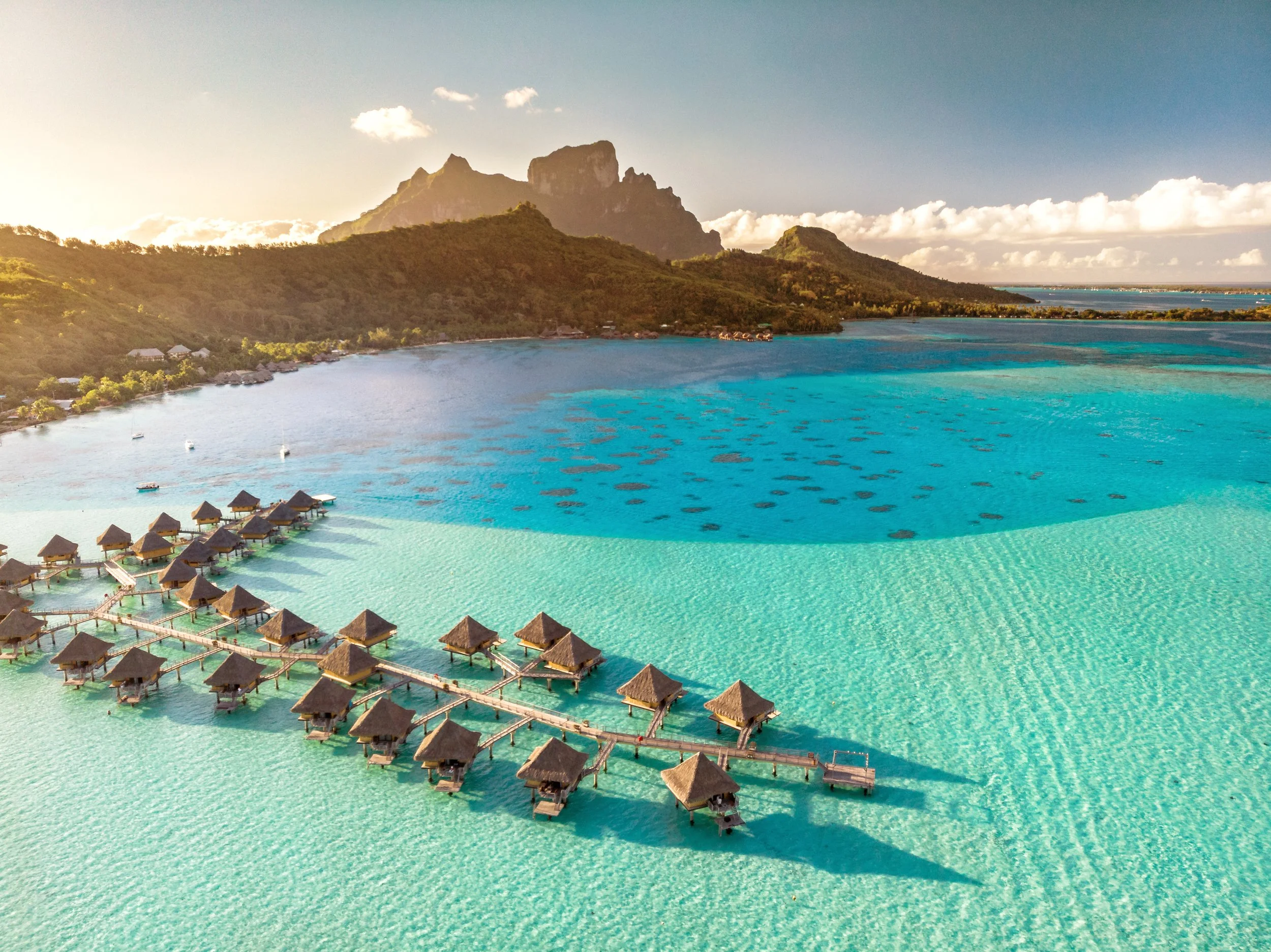 Overwater bungalows with thatched roofs on a turquoise lagoon, with a mountain range in the background under a partly cloudy sky during sunset.