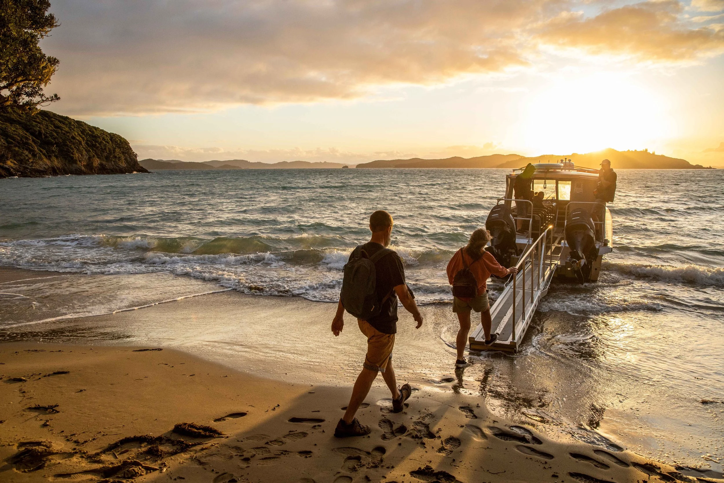 Two people board a boat at sunset in the Bay of Islands, New Zealand, with golden light reflecting across the calm water
