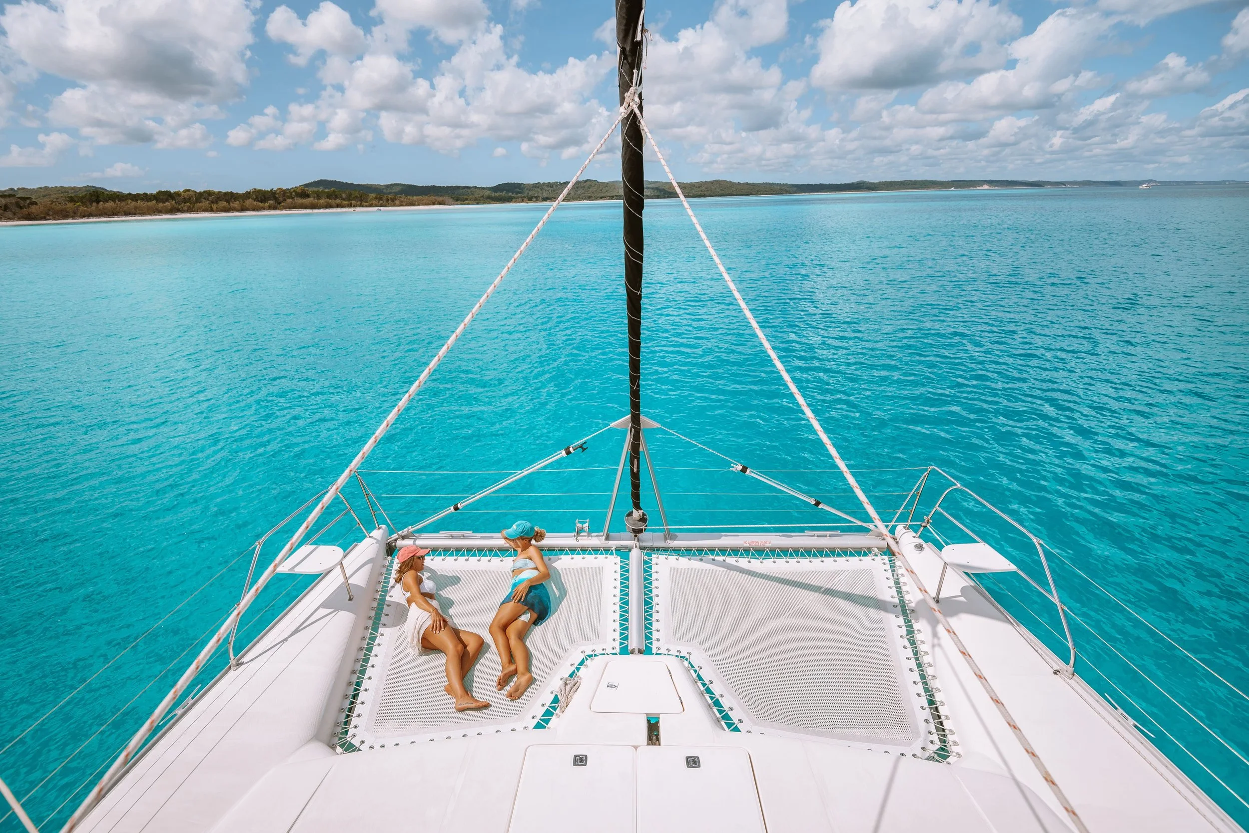 Two people relax on a luxury catamaran's netting in Australia, with turquoise water, a distant coastline, and blue skies with scattered clouds