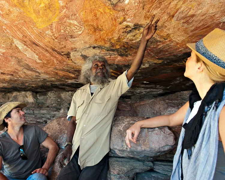 Three people in a cave with Aboriginal rock art, the older man with gray hair and a beard pointing at the wall, two women wearing hats looking at the rock art.