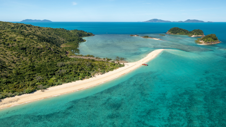A tropical beach with white sand and turquoise water, surrounded by green hills and small islands in the distance.