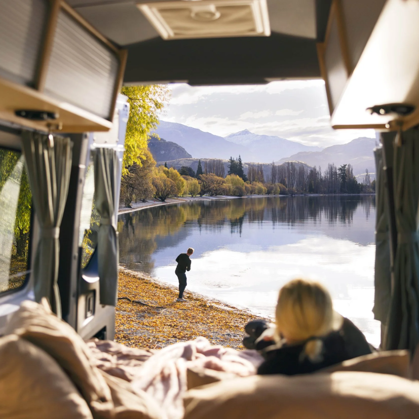 View of a woman from inside a camper van looking out at her boyfriend, who is skipping rocks on Lake Wanaka in Otago, New Zealand.
