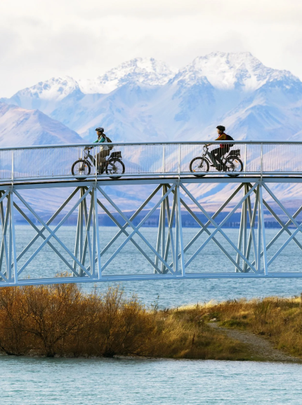 Two people riding bicycles on a high bridge over a lake with mountains in the background.