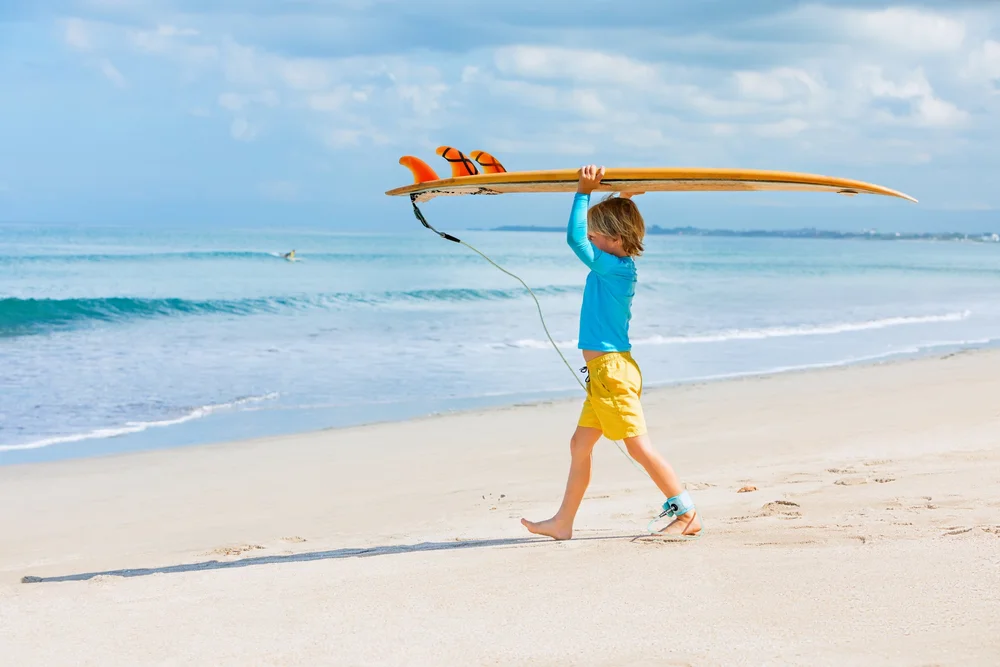 A happy young boy learns to surf on gentle waves during a kids’ surfing camp on the Gold Coast, Queensland, Australia, enjoying a fun summer vacation and active family lifestyle by the ocean.