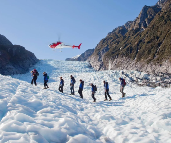 Hikers walk on snow and ice in a mountain valley as a helicopter flies overhead against a clear blue sky.