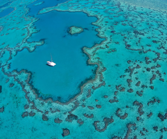 Aerial view of a sailboat anchored in a turquoise lagoon with coral reefs and shallow waters surrounding it.