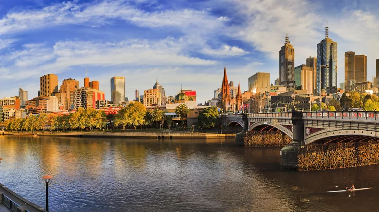 Skyline of Melbourne with river in foreground and paddleboarder on water, modern skyscrapers, historical buildings, and a bridge over the river during sunset.