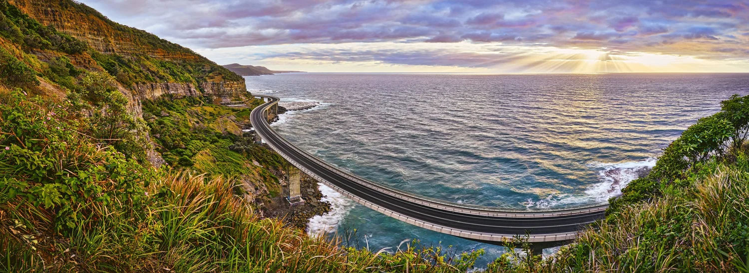 Panoramic view of the Sea Cliff Bridge in Wollongong, Australia, curving along the coastline above the ocean with cliffs and greenery in the background.