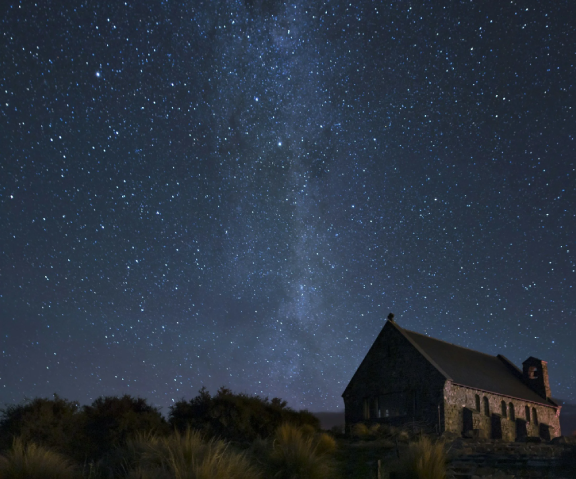 Night sky with numerous stars and the Milky Way galaxy visible above a dark, rustic stone house with a steep roof, surrounded by shrubs and low vegetation.