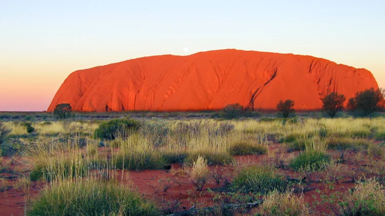 A large, red sandstone rock formation known as Uluru or Ayers Rock in the Australian Outback during sunset, with sparse desert vegetation and a small moon in the sky.