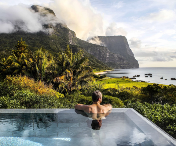Man relaxing in an infinity pool overlooking lush greenery, mountains, and ocean on a cloudy day.