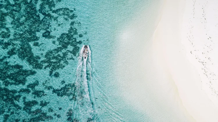 An aerial view of a boat moving across clear turquoise water near a white sandy beach.
