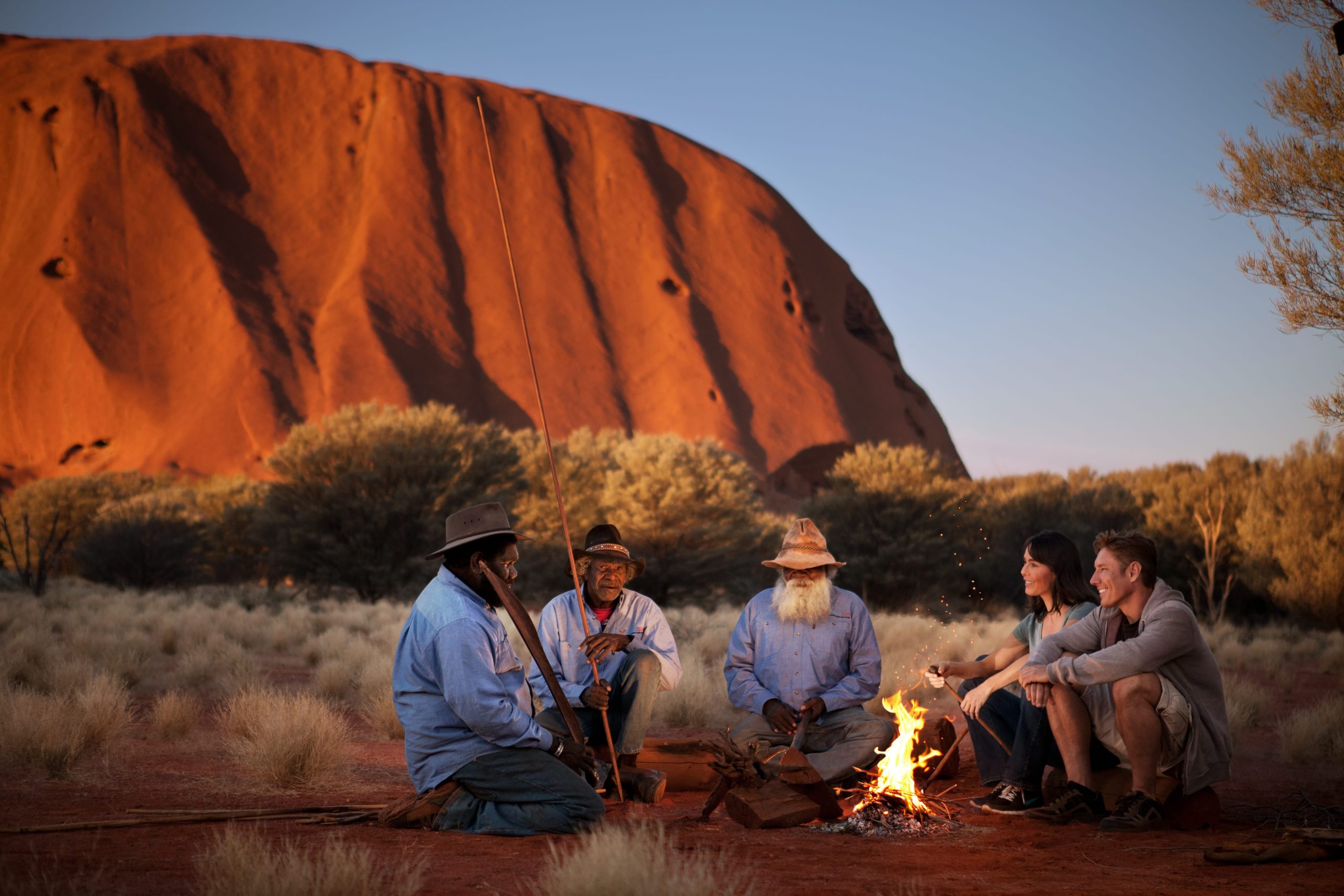 A group of Anangu elders shares stories around a fire with two travelers. In the background, Uluru stands against the blue sky.
