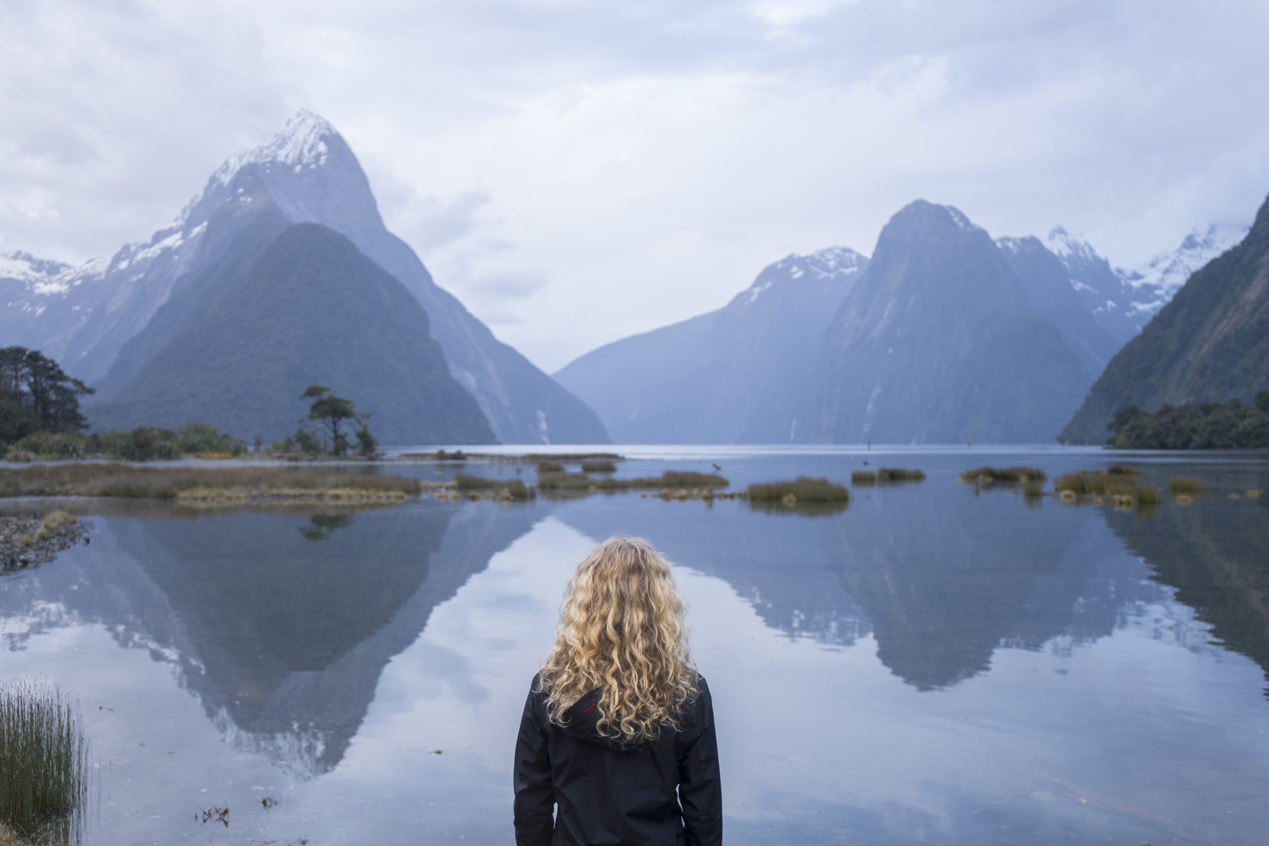 A woman looks out at landscape of Milford Sound. A mountain range rises from the water in the distance.