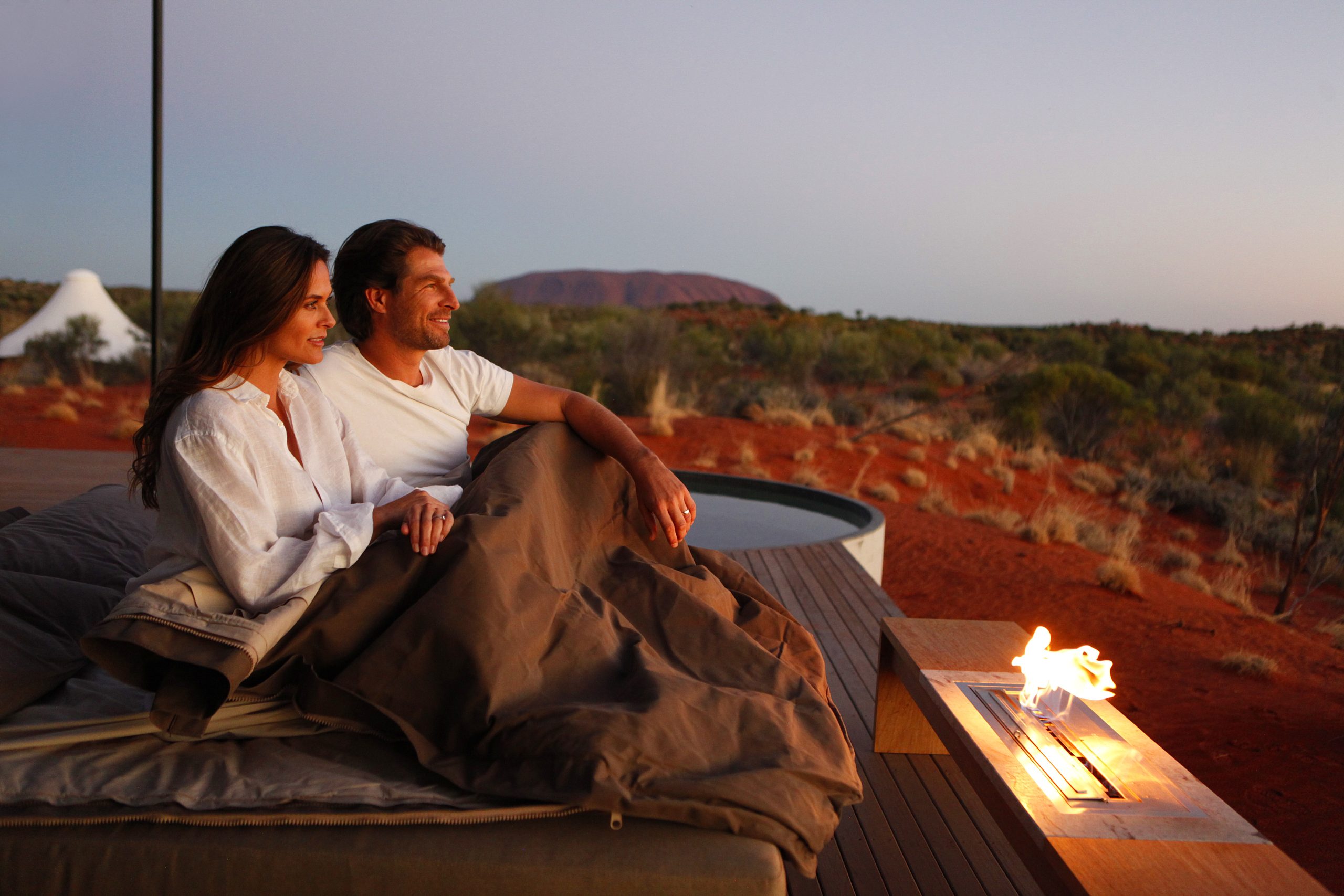 A couple sitting on the deck of their luxury Uluru campsite, enjoying the view at sunset near a fire.